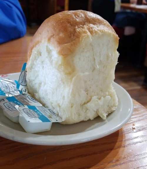 A large, fluffy bread roll on a plate next to small packets of butter.