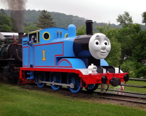 A blue cartoon train with a smiling face, named Thomas, on a track surrounded by greenery.