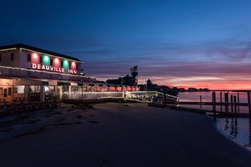 Deauville Inn illuminated at sunset, with a sandy beach and calm water reflecting the colorful sky.