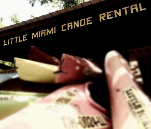 Sign for "Little Miami Canoe Rental" with colorful kayaks in the foreground, slightly blurred.