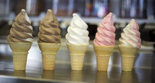Five ice cream cones with swirled soft serve in chocolate, vanilla, and strawberry flavors, lined up on a counter.