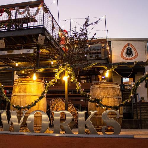 A festive outdoor space with barrels, string lights, and holiday decorations, featuring a sign that reads "WORKS."