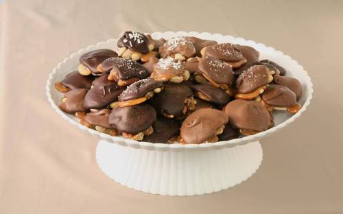 A white pedestal dish filled with assorted chocolate-covered treats, including nuts and caramel, on a beige background.