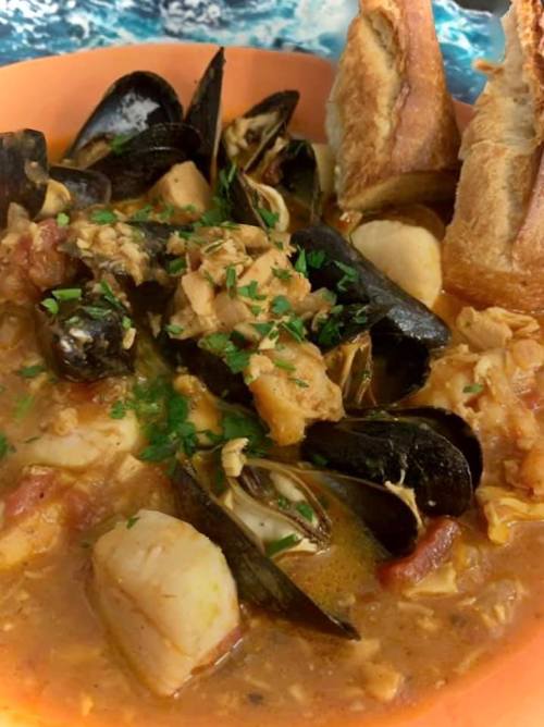A bowl of seafood stew with mussels, scallops, and herbs, served with slices of crusty bread on the side.