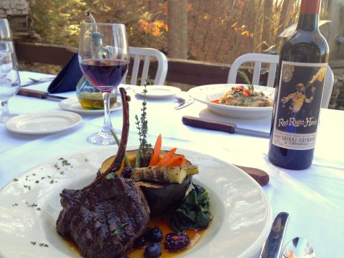 A gourmet meal featuring lamb chops, vegetables, and a bottle of red wine on a beautifully set table outdoors.
