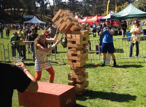 A woman in a sparkly dress attempts to balance a tall Jenga tower at an outdoor event with spectators in the background.