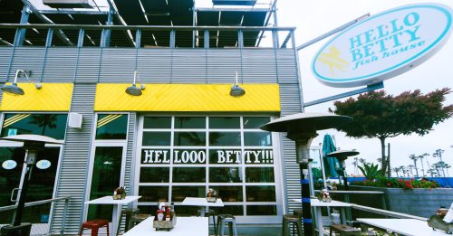 Exterior of Hello Betty Fish House with bright yellow accents and outdoor seating, featuring a playful sign.
