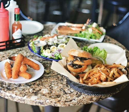 A table with a burger and fries, mozzarella sticks, and a fresh salad with sushi in the background.
