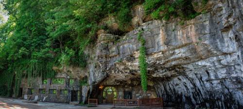A rocky cliffside with lush greenery and a stone structure built into the cave, featuring large windows and a wooden deck.