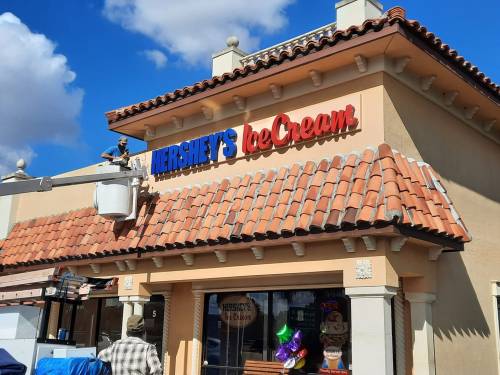 A worker on a lift adjusts the sign for Hershey's Ice Cream on a building with a tiled roof under a blue sky.
