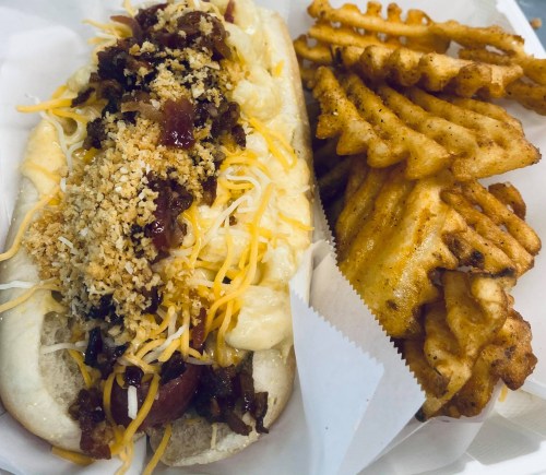 A loaded hot dog topped with cheese, bacon, and breadcrumbs, served with a side of crispy waffle fries.