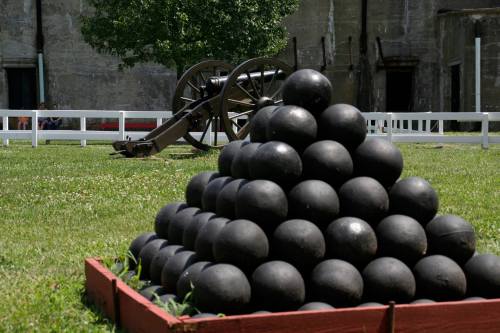 A pyramid of cannonballs in a grassy area, with a cannon and historic building in the background.