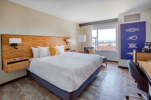 Modern hotel room featuring a queen bed, desk, and large window with a view. Warm tones and stylish decor.