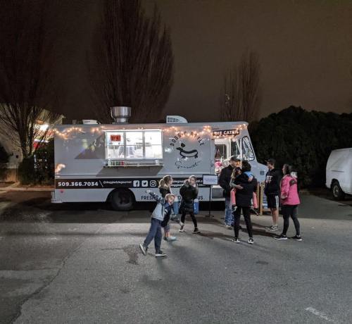 A food truck decorated with lights at night, with a group of children gathered around it in a parking lot.