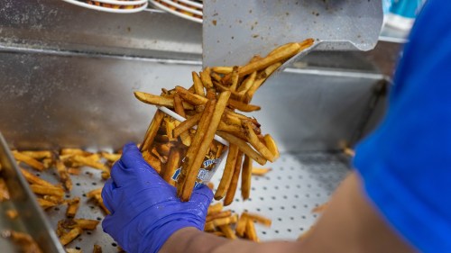 A hand in a blue glove pours crispy French fries into a container from a metal tray.
