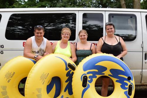 Four people stand in front of a van, holding yellow inflatable tubes, smiling and enjoying a sunny day outdoors.