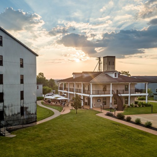 A scenic view of a building with a porch, surrounded by green grass, under a sunset sky with clouds.