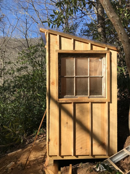 A small wooden shed with a window, surrounded by trees and nature, on a sunny day.