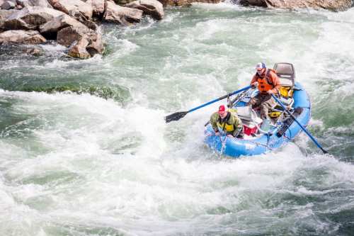 Two people navigate a blue raft through turbulent whitewater rapids, surrounded by rocky banks.