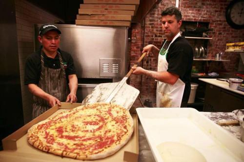Two chefs in a pizzeria, one placing a large pizza in a box while the other holds a pizza peel.