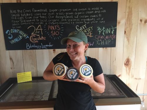 A woman holds three tubs of ice cream in front of a menu board at a farm, smiling and wearing a cap.