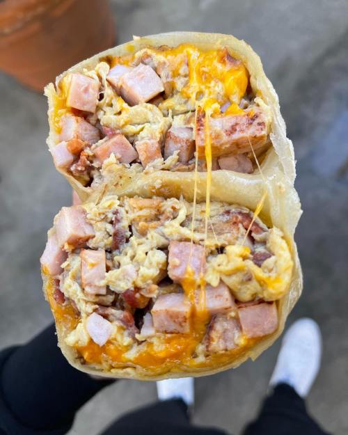 A close-up of a halved burrito filled with ham, scrambled eggs, and melted cheese, held in hands.