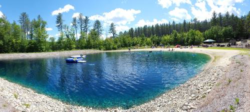 A serene pond surrounded by trees, with people swimming and relaxing on the shore under a bright blue sky.