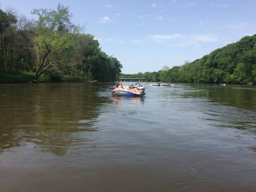 A group of people in a raft on a calm river, surrounded by lush green trees under a clear blue sky.