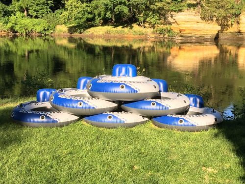 A stack of blue and gray inflatable tubes on green grass by a calm river surrounded by trees.