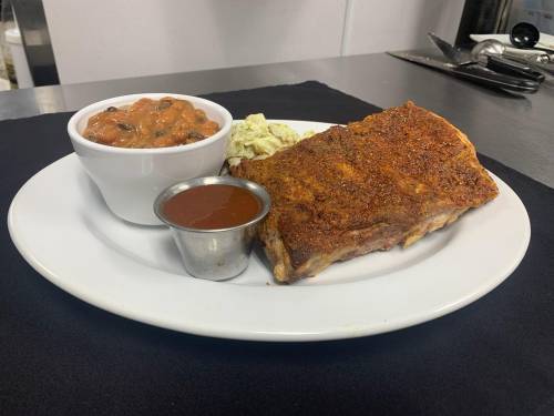A plate featuring seasoned ribs, a side of baked beans, and coleslaw, with barbecue sauce in a small cup.