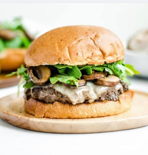 A juicy burger with melted cheese, mushrooms, and fresh greens on a toasted bun, served on a wooden plate.