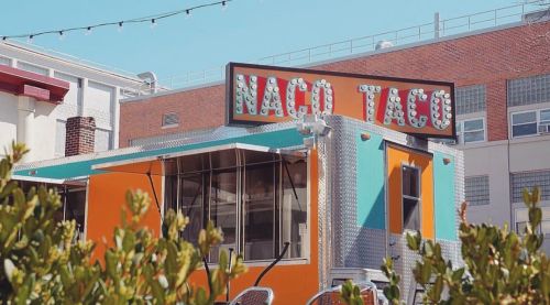 A colorful taco food truck with a bright sign, surrounded by greenery and buildings in the background.