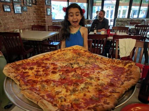 A surprised girl stands in front of an enormous pizza, showcasing its large size and toppings in a restaurant setting.