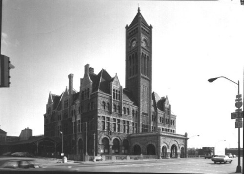 Historic clock tower building with intricate architecture, set on a street corner in black and white.