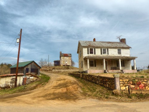 A weathered farmhouse and outbuilding on a dirt road, surrounded by trees under a cloudy sky.