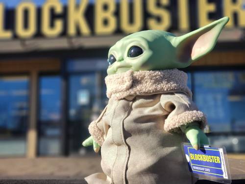 A toy figure resembling a green alien stands outside a Blockbuster store, holding a membership card.