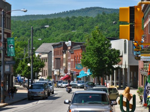 A scenic street view with shops, trees, and mountains in the background on a sunny day. Cars are parked along the road.