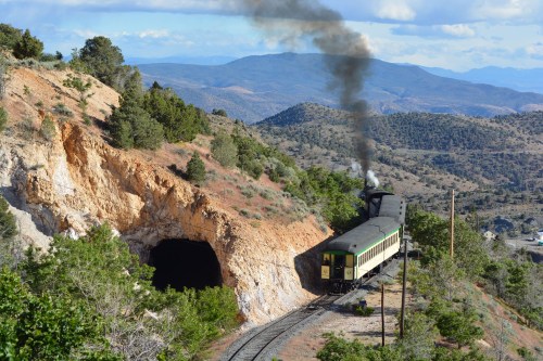 A vintage steam train travels along a mountain track near a tunnel, surrounded by lush greenery and distant hills.
