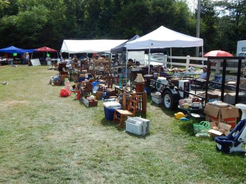 A bustling outdoor market with various tents displaying furniture, antiques, and household items on a grassy field.