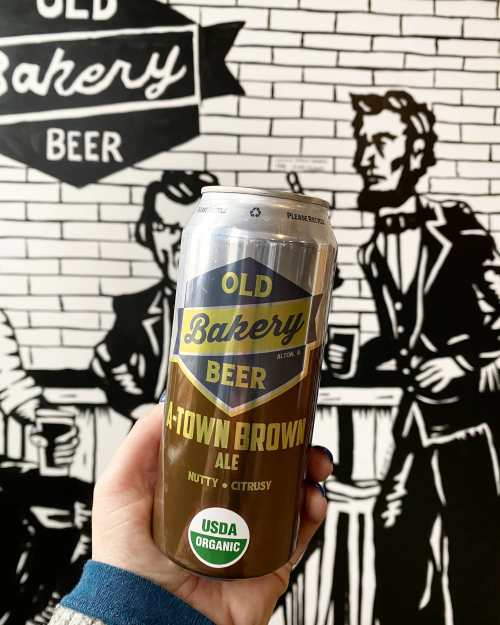 A hand holding a can of Old Bakery Beer A-Town Brown Ale in front of a mural featuring vintage-style characters.