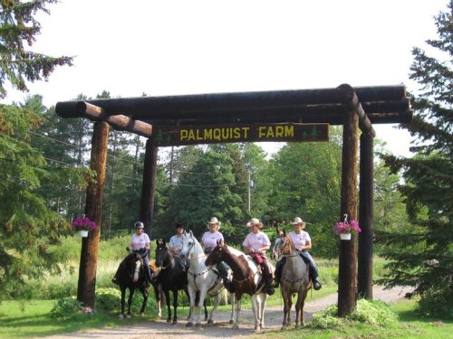 Four riders on horseback stand under a wooden archway labeled "Palmquist Farm," surrounded by trees and greenery.