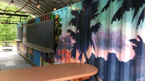 Colorful mural of palm trees and sunset on shipping container walls, with a wooden bar in a rustic outdoor setting.