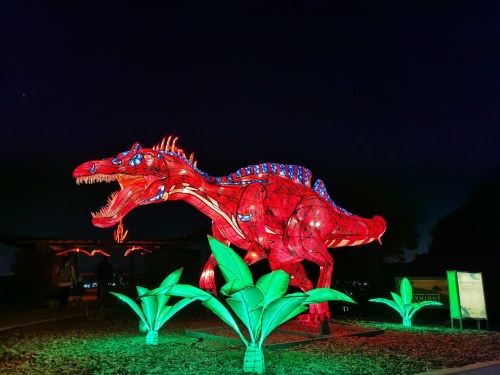 A large, illuminated red dinosaur sculpture stands among green lighted plants at night.