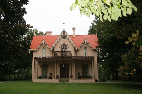 A pink Gothic-style house with a red roof, surrounded by greenery and trees.