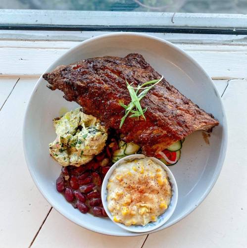 A plate featuring grilled ribs, potato salad, corn, and a side of beans, garnished with fresh herbs.