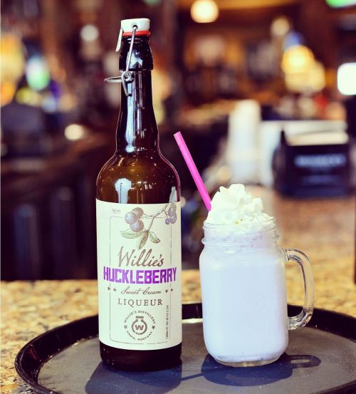 A bottle of Willie's Huckleberry liqueur next to a frosty drink topped with whipped cream and a pink straw.