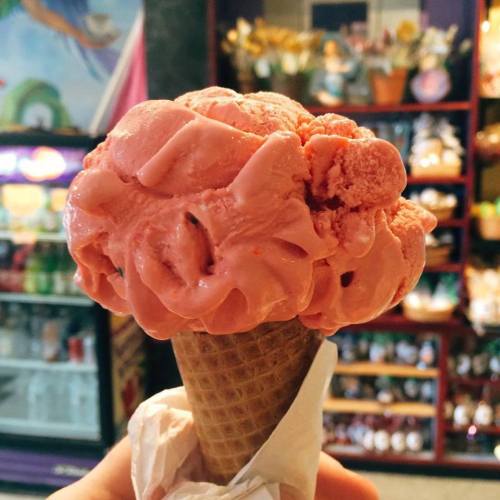 A close-up of a large scoop of pink ice cream on a waffle cone, held in a hand, with a colorful background.