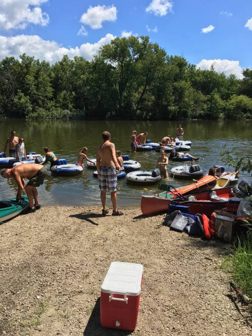 A group of people in inner tubes enjoying a sunny day on a river, with kayaks and a cooler on the shore.