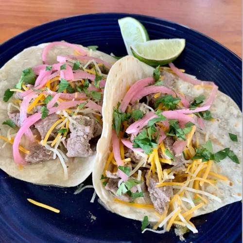 Two tacos filled with beef, cheese, pink onions, and cilantro, served with a lime wedge on a blue plate.