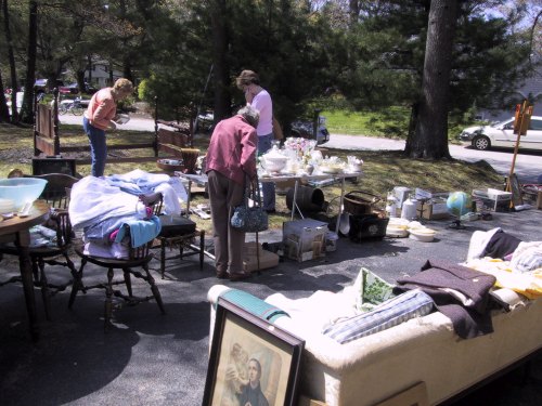 A yard sale scene with people browsing items like furniture, clothing, and household goods among trees.